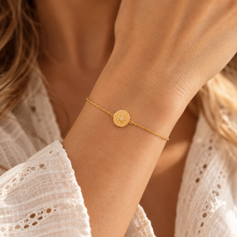 Bracelet Dalia porté au poignet, chaîne fine dorée et médaillon rond strié orné d’une étoile et de pierres blanches, blouse écrue et lumière naturelle.