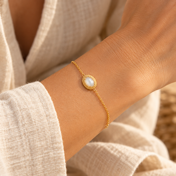 Bracelet Elyssia porté au poignet, chaîne fine dorée et pierre de lune ovale entourée d’un motif doré tressé, blouse écrue et lumière naturelle.