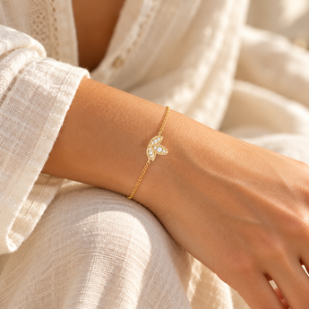 Bracelet Jalina porté au poignet, chaîne fine dorée et motif à trois feuilles serties de pierres blanches, vêtement écru et lumière naturelle.