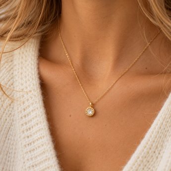 Collier Auréa porté, chaîne fine dorée et petit pendentif rond à pierre claire facettée entourée de détails dorés, maille écrue et lumière naturelle.